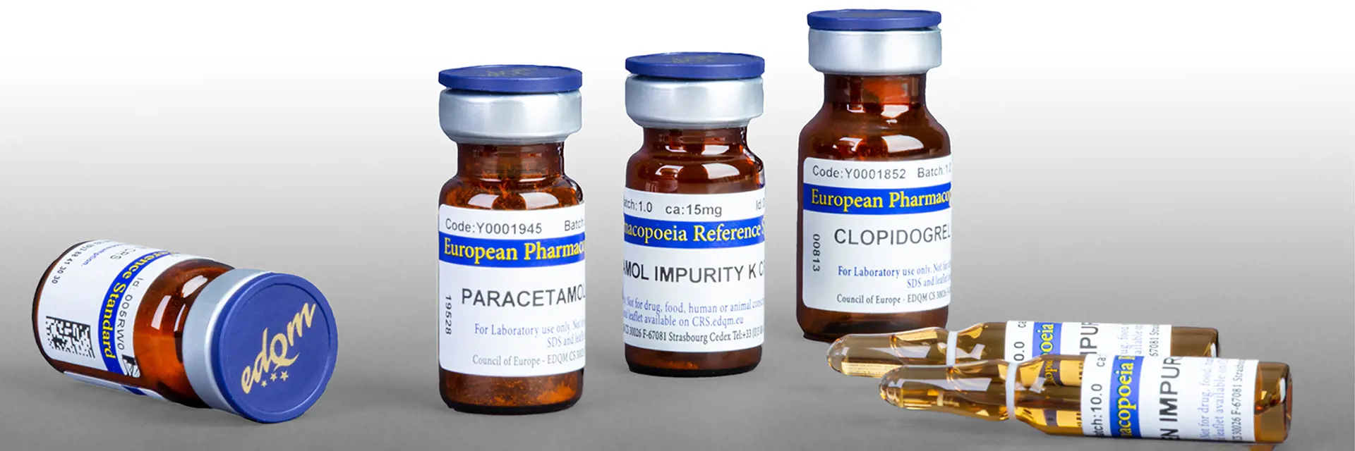 Brown glass vials and ampoules containing primary reference standards from the European Pharmacopoeia. The caps are blue with EDQM in gold print and the labels are white, blue and yellow.